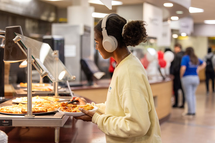 student with white headphones in a yellow sweatshirt putting food on her plate in the cafeteria